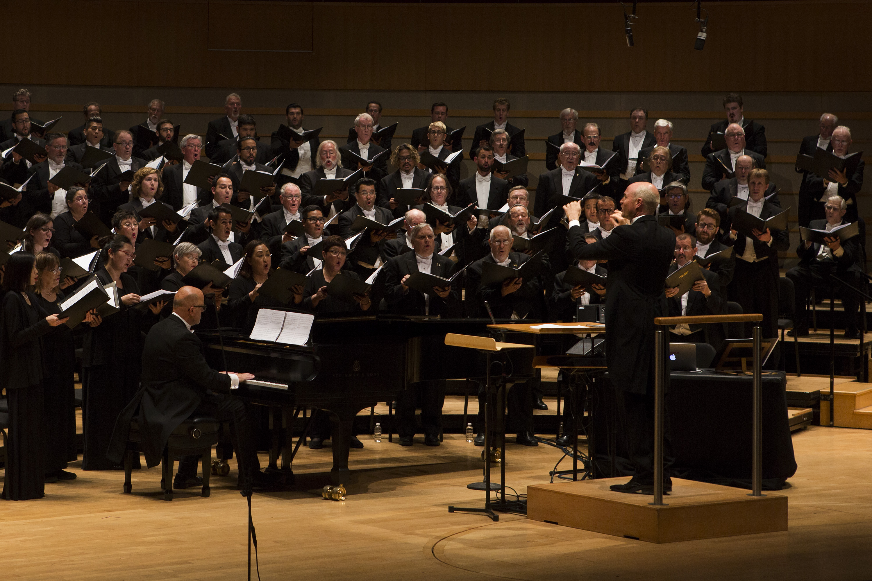The Pacific Chorale of Orange County performs on stage, accompanied by a pianist. The conductor leads the ensemble as singers hold sheet music under bright lights in a concert hall setting.