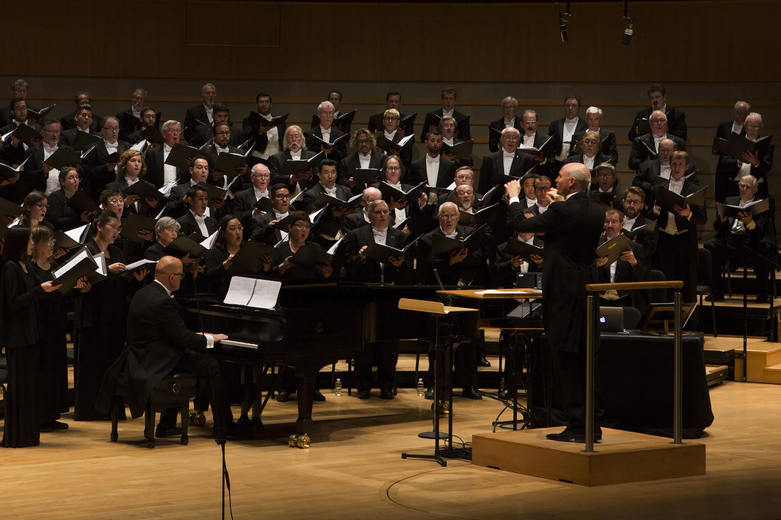 The Pacific Chorale of Orange County performs on stage, accompanied by a pianist. The conductor leads the ensemble as singers hold sheet music under bright lights in a concert hall setting.