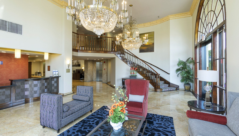 Spacious Best Western Costa Mesa hotel lobby with marble floors, elegant chandeliers, a grand staircase, modern sofas, a glass coffee table with flowers, and large arched windows letting in natural light.