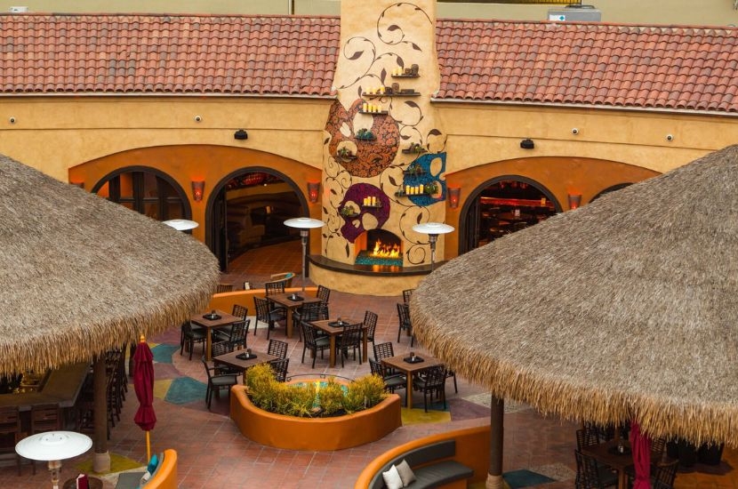 Outdoor restaurant patio at Triangle Square Costa Mesa, featuring thatched-roof umbrellas, empty tables and chairs, and a decorative wall with colorful artistic fish and candles above a fireplace in a warm, cozy setting.