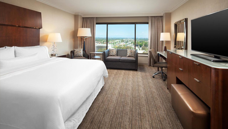 Modern hotel room at Westin South Coast Plaza with a large bed, flat-screen TV, wooden furniture, sofa, desk and chair, lamps, and a large window offering a stunning city view with blue sky and greenery outside.
