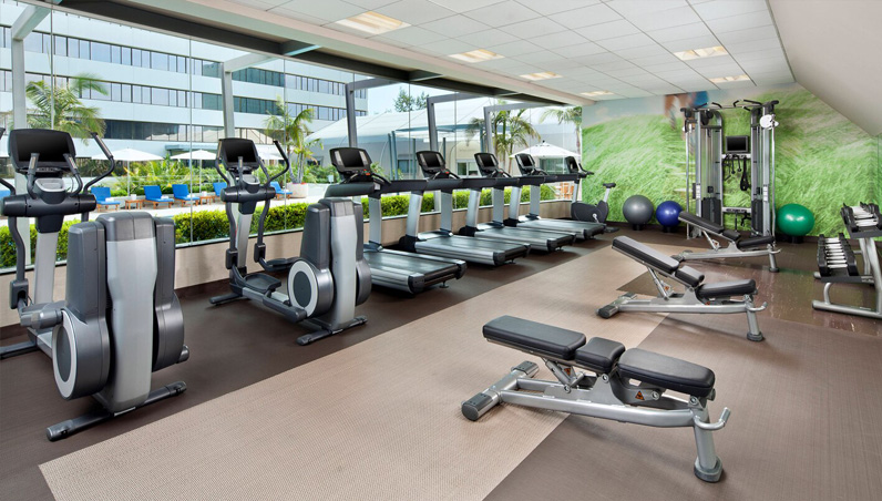A modern gym at Westin South Coast Plaza with treadmills, elliptical machines, weight benches, and strength training equipment, featuring large windows overlooking the pool and office buildings.