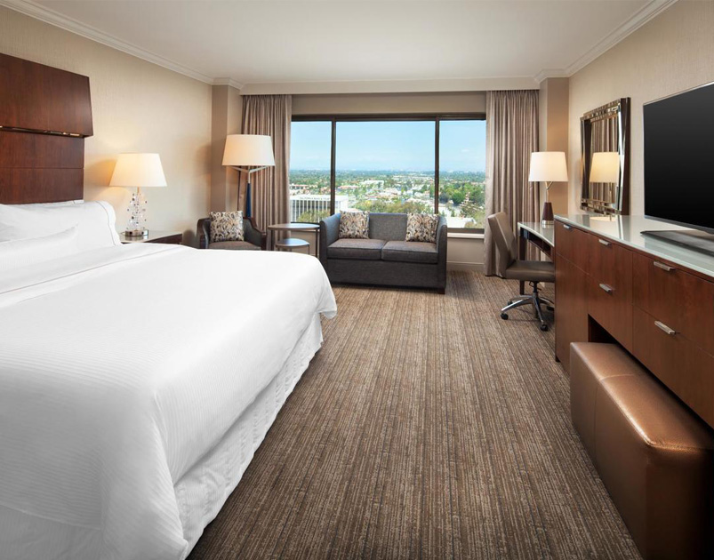 Modern hotel room at Westin South Coast Plaza with a large bed, two side tables with lamps, a sofa by a window offering city views, a desk and chair, flat-screen TV, and neutral-colored decor.
