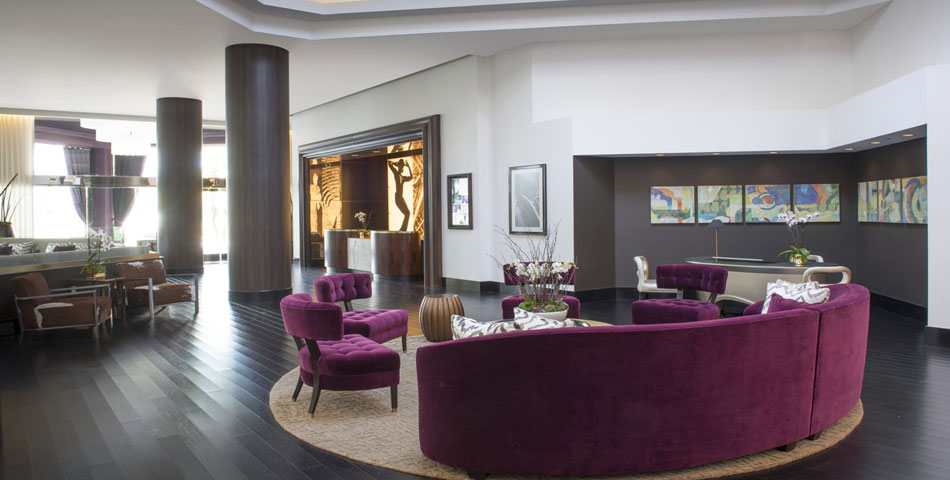 A modern hotel lobby with dark wood floors, a round beige rug, purple velvet sofas and chairs, art on the walls, and large windows letting in natural light.