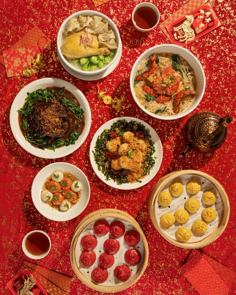 A festive table with various Chinese dishes, including dumplings, noodles, greens, and soup, set on a red, gold-speckled tablecloth with tea cups, a teapot, and traditional red envelopes for Lunar New Year.