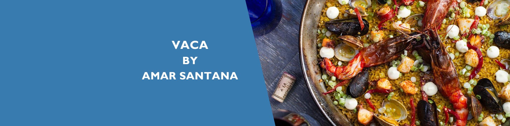A pan of colorful seafood paella with shrimp, mussels, and vegetables sits on a table at Vaca Restaurant Costa Mesa, beside a cork and glass, next to a blue background with the text “Vaca by Amar Santana.”.