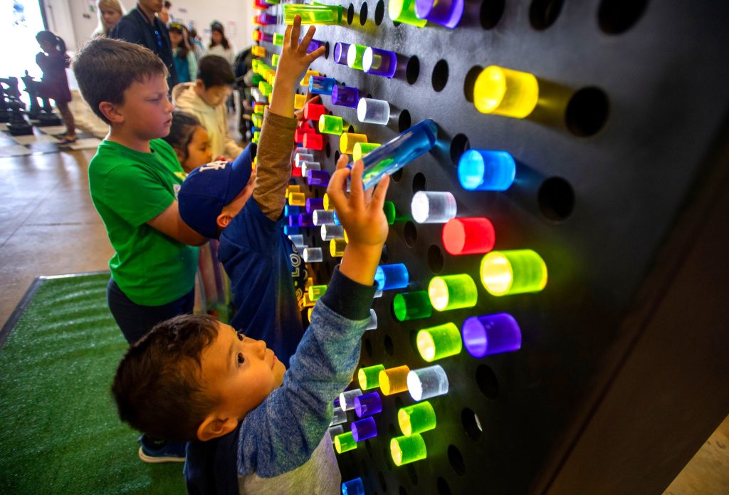 Three young children are placing large, colorful plastic pegs into a black, wall-mounted board with holes at an interactive exhibit. Other people and activity can be seen in the background.