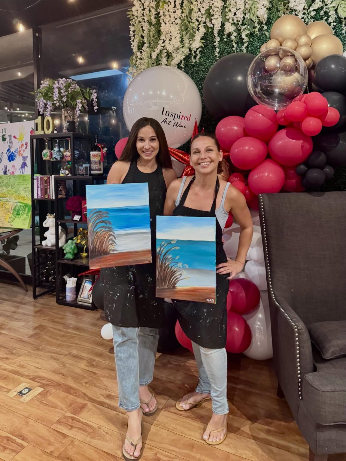 Two women smile and stand side by side, each holding a beach-themed painting. They wear aprons and casual clothes. Behind them are colorful balloons, flowers, and a “Inspired Art” sign, creating a festive atmosphere.