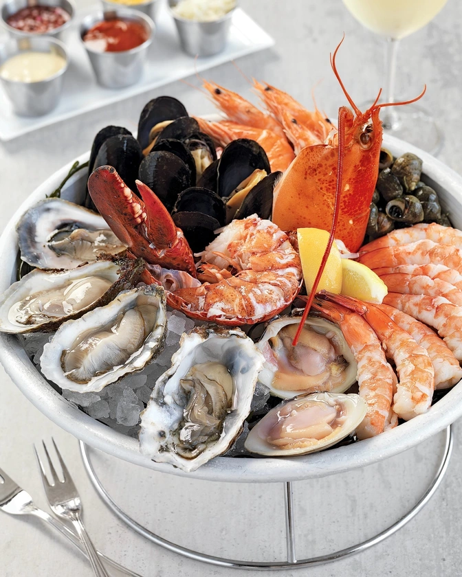 A seafood platter on ice featuring oysters, mussels, shrimp, prawns, lobster claw, and lemon wedges, served in a metal bowl with sauces in the background.