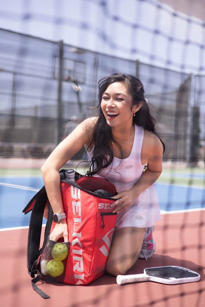 A smiling woman in athletic wear kneels on a tennis court, placing pickleballs and gear into a red Selkirk sports bag. A paddle and water bottle are on the ground beside her. The scene is viewed through a net.
