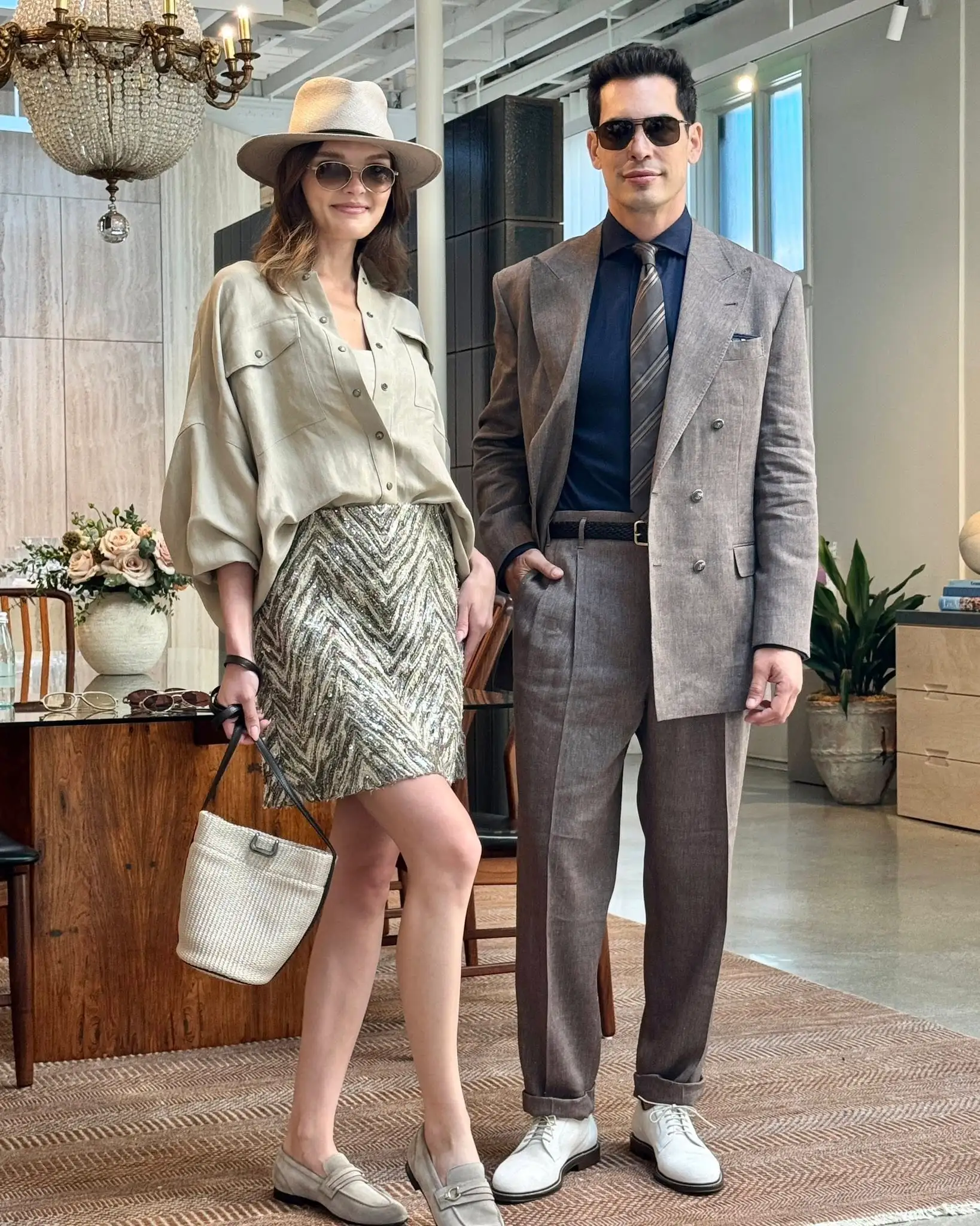 A stylish woman in a beige blouse, patterned skirt, hat, and sunglasses stands next to a man in a gray suit, black shirt, tie, and sunglasses. They pose indoors by a dining table with floral decor and natural light.