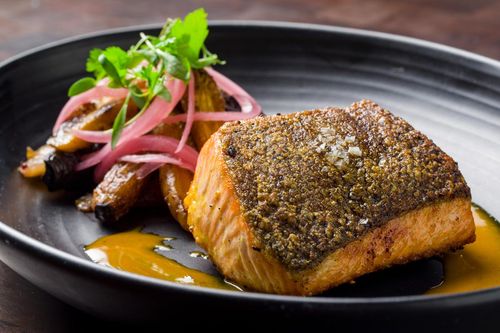 A plated dish featuring a seared, crispy-skinned salmon fillet, served with roasted vegetables, pickled red onions, fresh herbs, and a pool of orange sauce on a black plate.