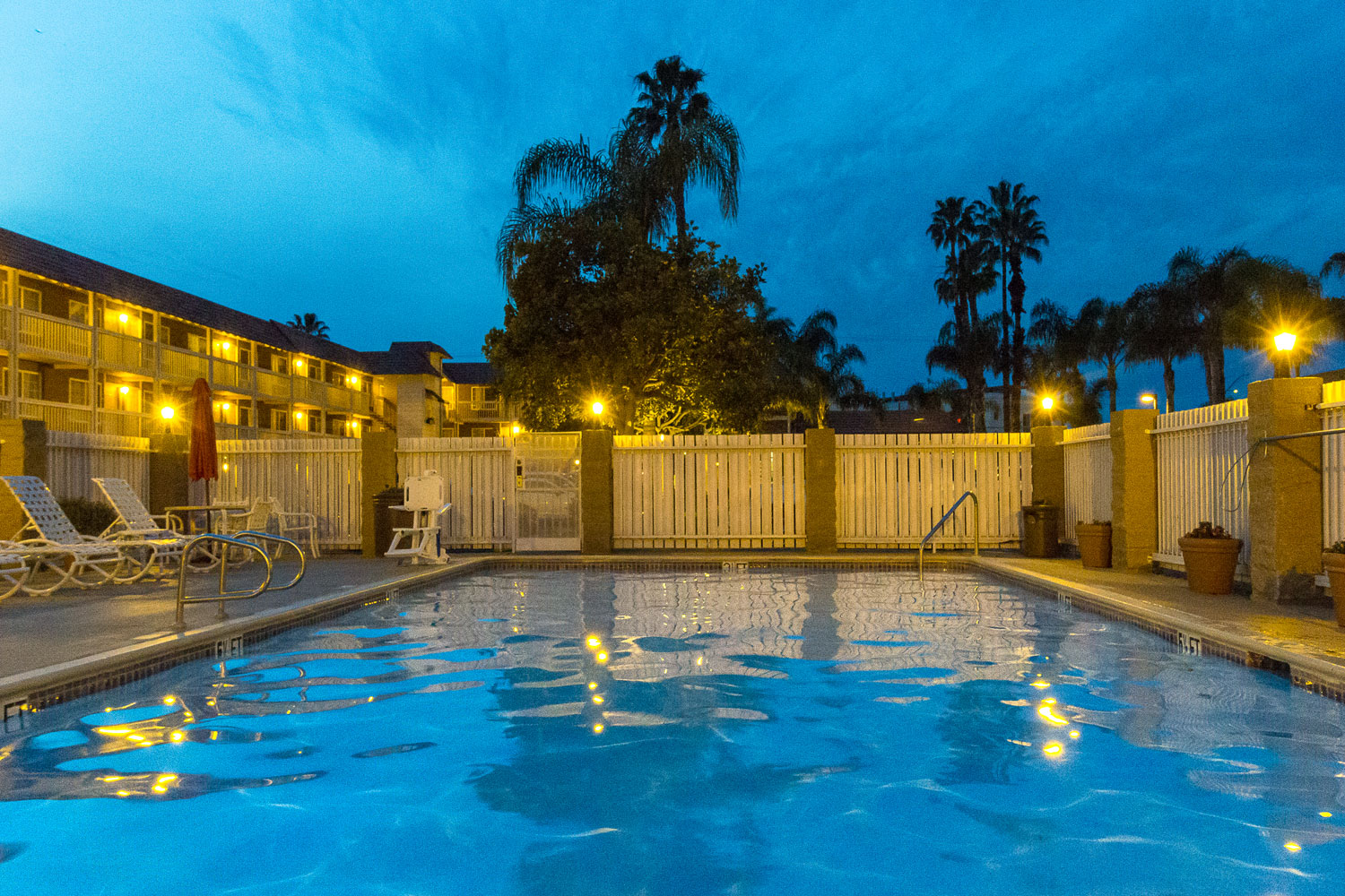 A swimming pool at dusk at Ramada Costa Mesa, surrounded by lounge chairs and a white fence, with palm trees and illuminated hotel buildings in the background under a deep blue sky.