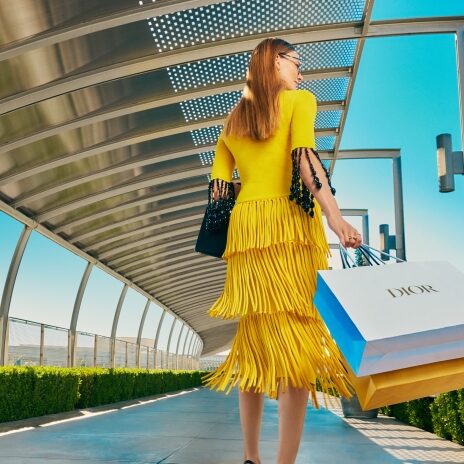 A woman in a bright yellow fringe dress and high heels walks outdoors on a covered walkway, carrying luxury shopping bags, including a large Dior bag, under a clear blue sky.