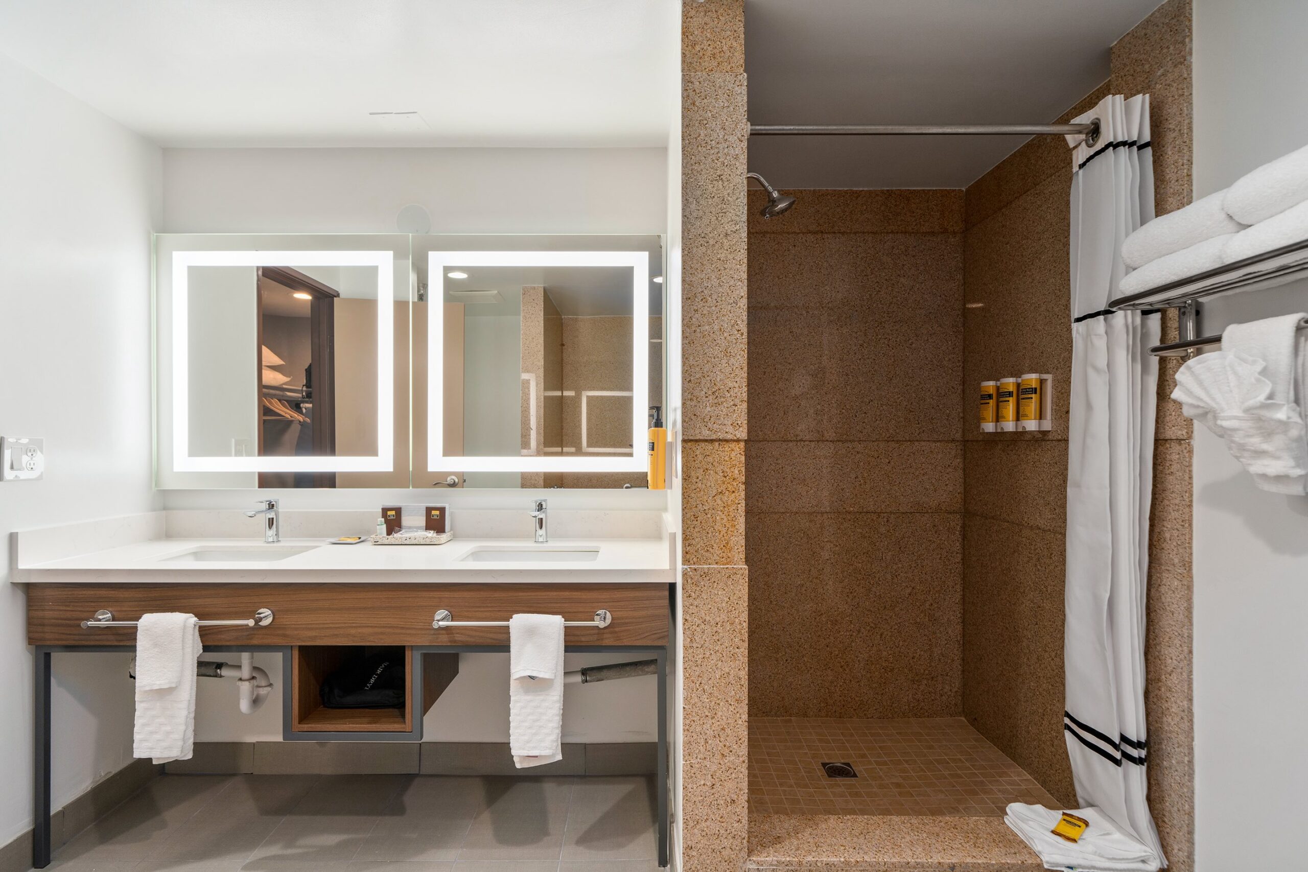 A modern bathroom at Best Western Costa Mesa features a double sink vanity, large illuminated mirrors, and a walk-in shower with beige tiles. Towels hang neatly by the sink and shower, while toiletries are arranged on convenient shelves.