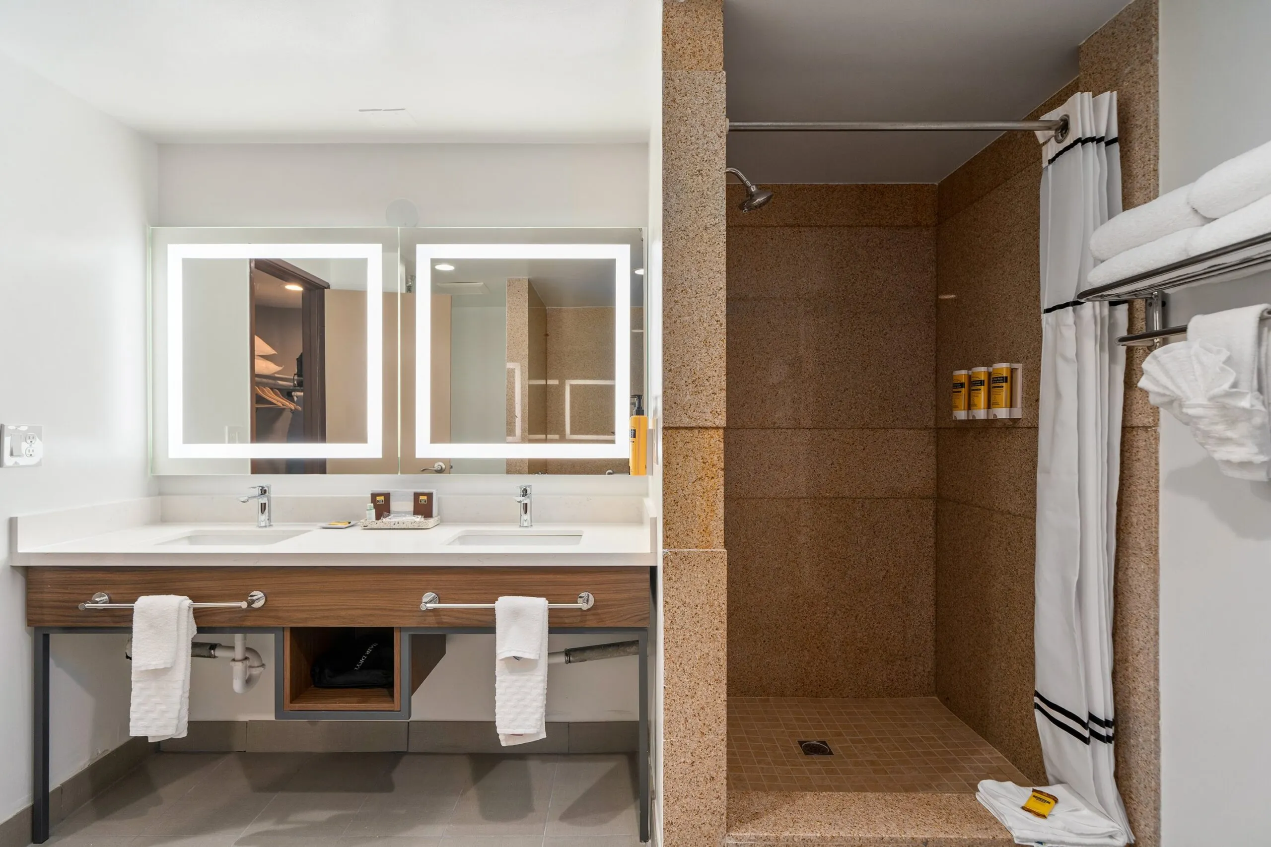 A modern bathroom at Best Western Costa Mesa features a double sink vanity, large illuminated mirrors, and a walk-in shower with beige tiles. Towels hang neatly by the sink and shower, while toiletries are arranged on convenient shelves.