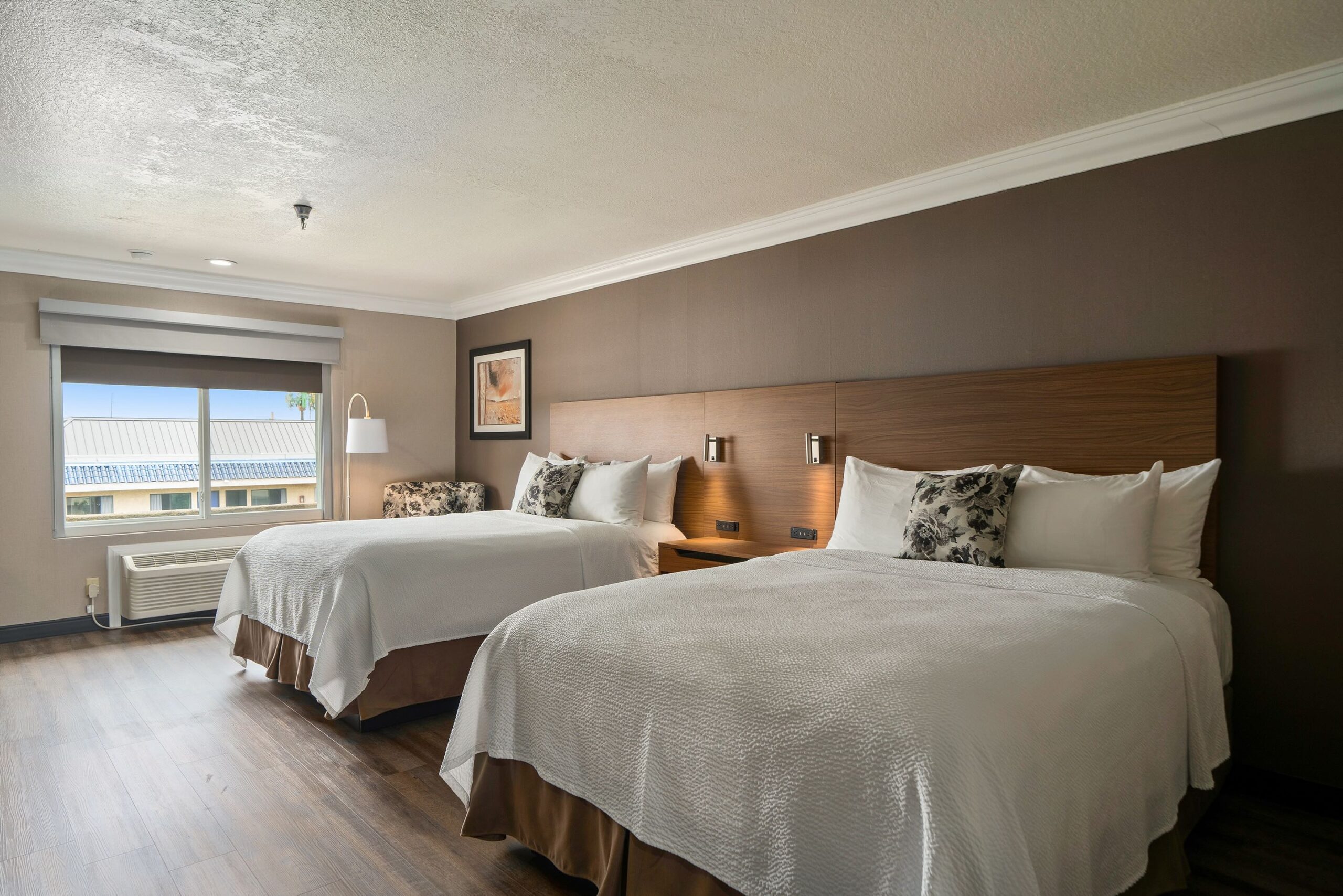 A hotel room at Best Western Costa Mesa with two neatly made double beds, floral accent pillows, a window with a view, an armchair, wall art, wooden floors, and a wall-mounted lamp.