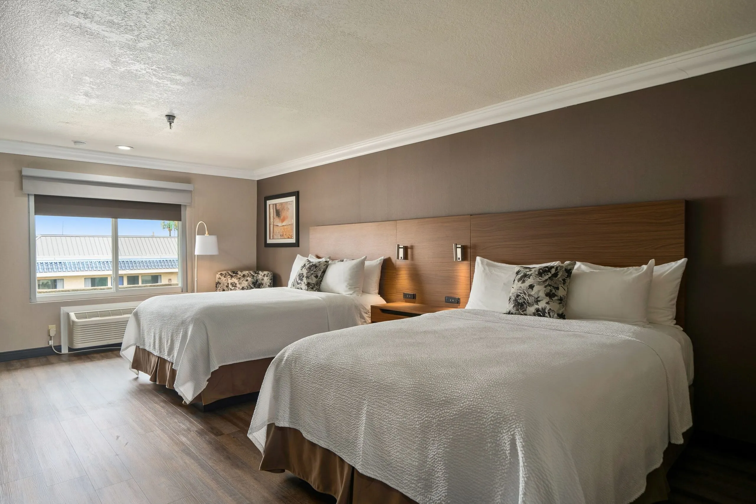 A hotel room at Best Western Costa Mesa with two neatly made double beds, floral accent pillows, a window with a view, an armchair, wall art, wooden floors, and a wall-mounted lamp.