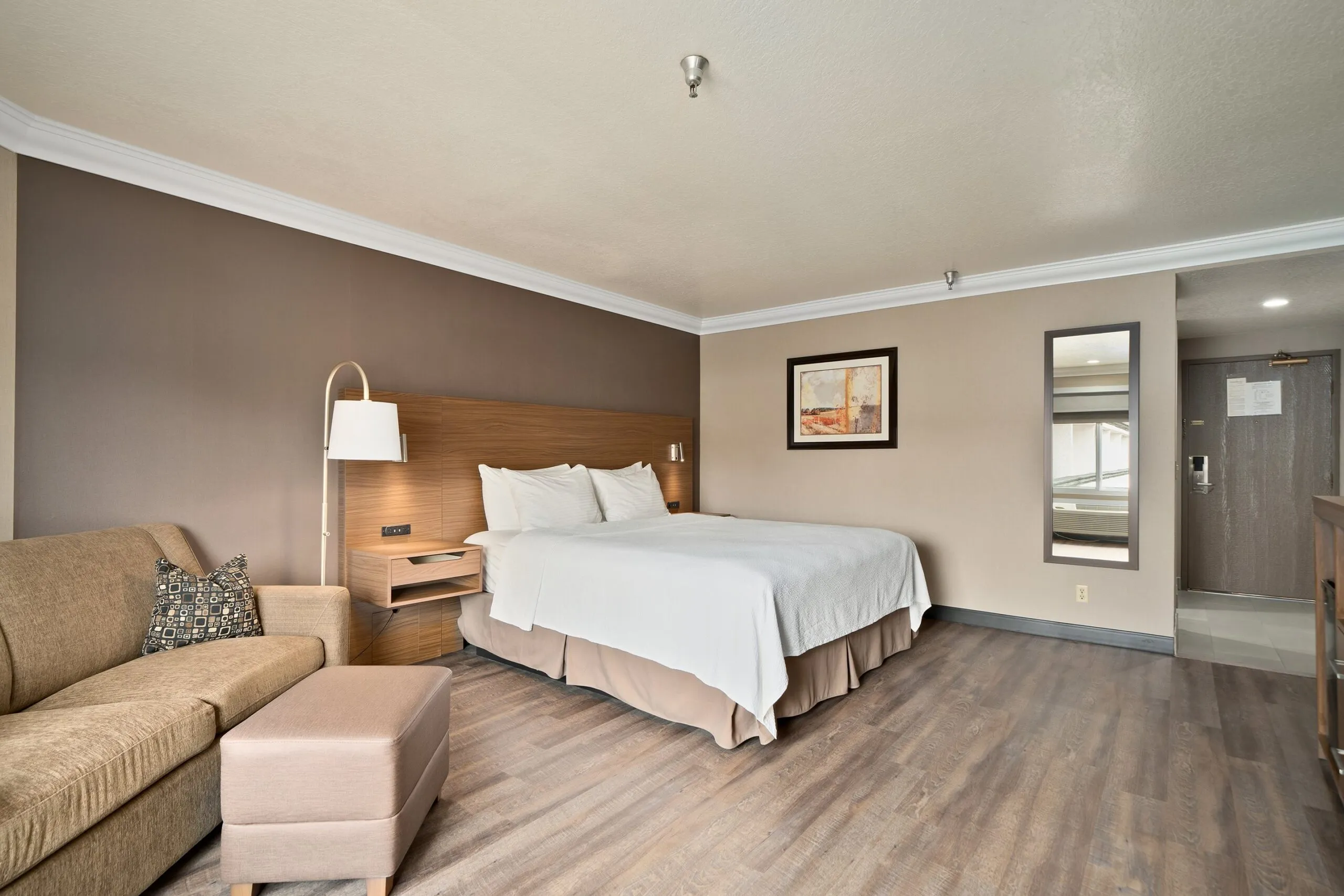 A modern Best Western Costa Mesa hotel room with a king bed, brown accent wall, nightstands, a tan sofa and ottoman, wall lamp, framed artwork, large mirror, wood-look flooring, and entryway with door visible in the background.