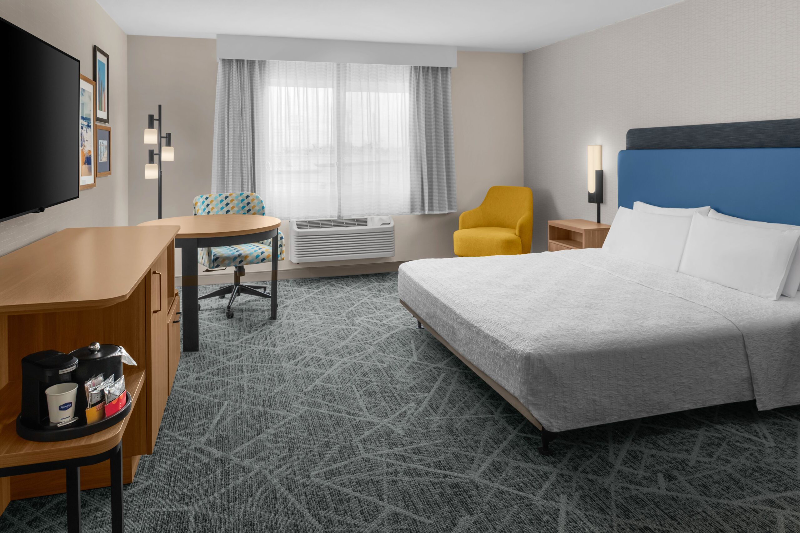 Modern hotel room at Hampton Inn Costa Mesa Newport Beach with a bed, blue headboard, yellow armchair, desk by the window, flat-screen TV, wooden furniture, coffee maker on a cabinet, light grey walls, and patterned grey carpet.