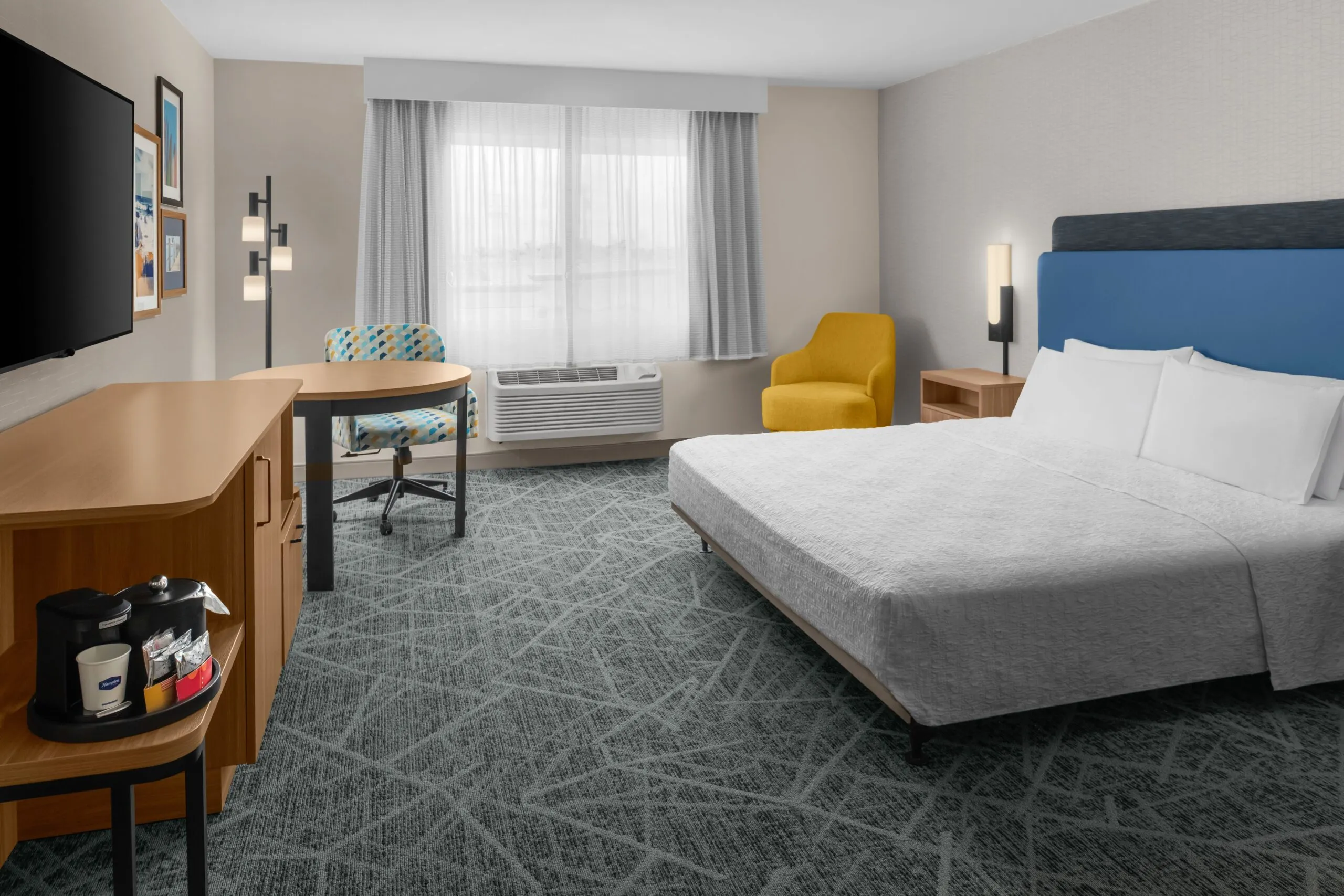 Modern hotel room at Hampton Inn Costa Mesa Newport Beach with a bed, blue headboard, yellow armchair, desk by the window, flat-screen TV, wooden furniture, coffee maker on a cabinet, light grey walls, and patterned grey carpet.