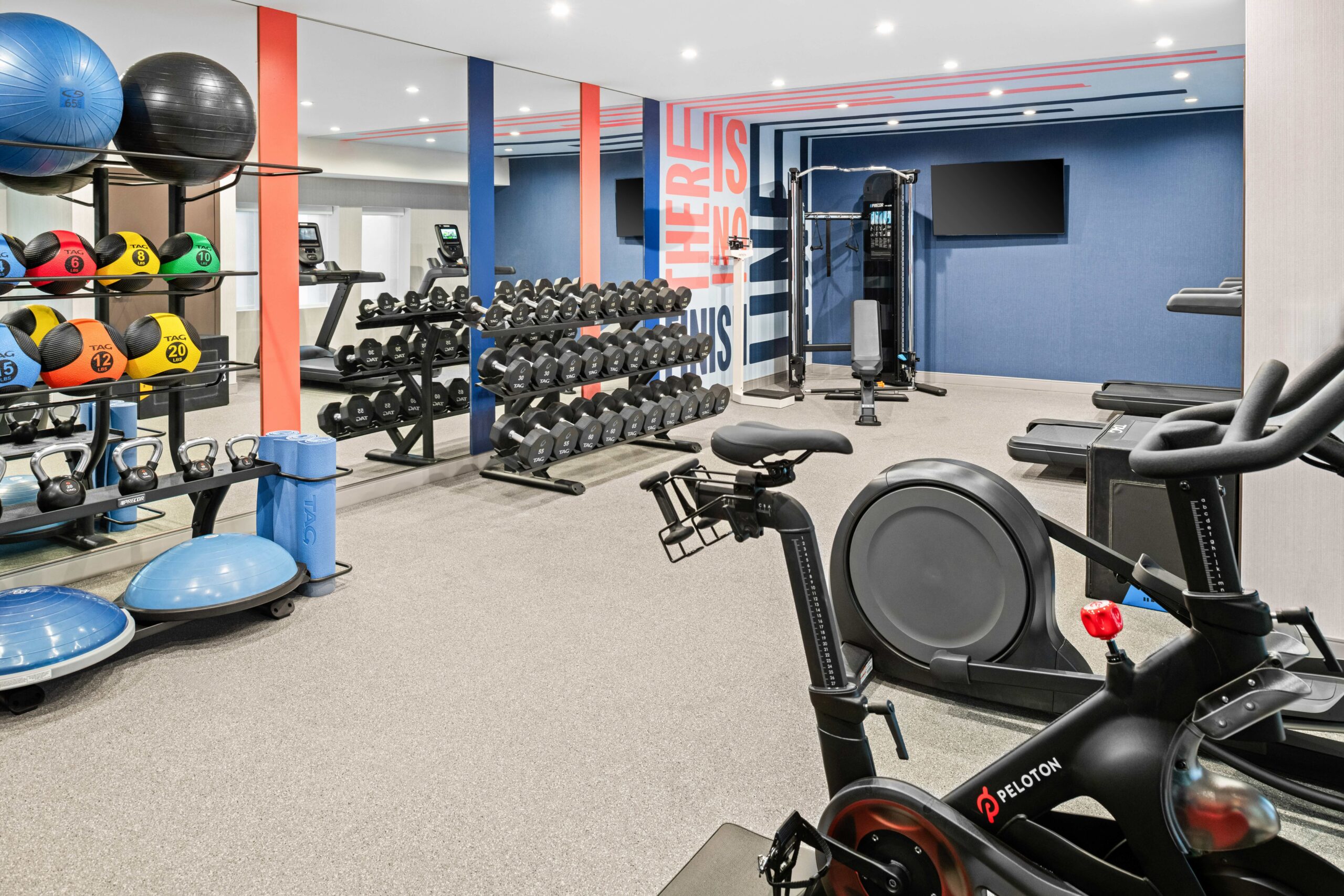 Modern gym at Hampton Inn Costa Mesa Newport Beach with exercise bikes, free weights, dumbbells, kettlebells, medicine balls, mats, BOSU balls, resistance bands, and cardio machines. Blue, gray, and red accents with bright lighting and a wall-mounted TV.