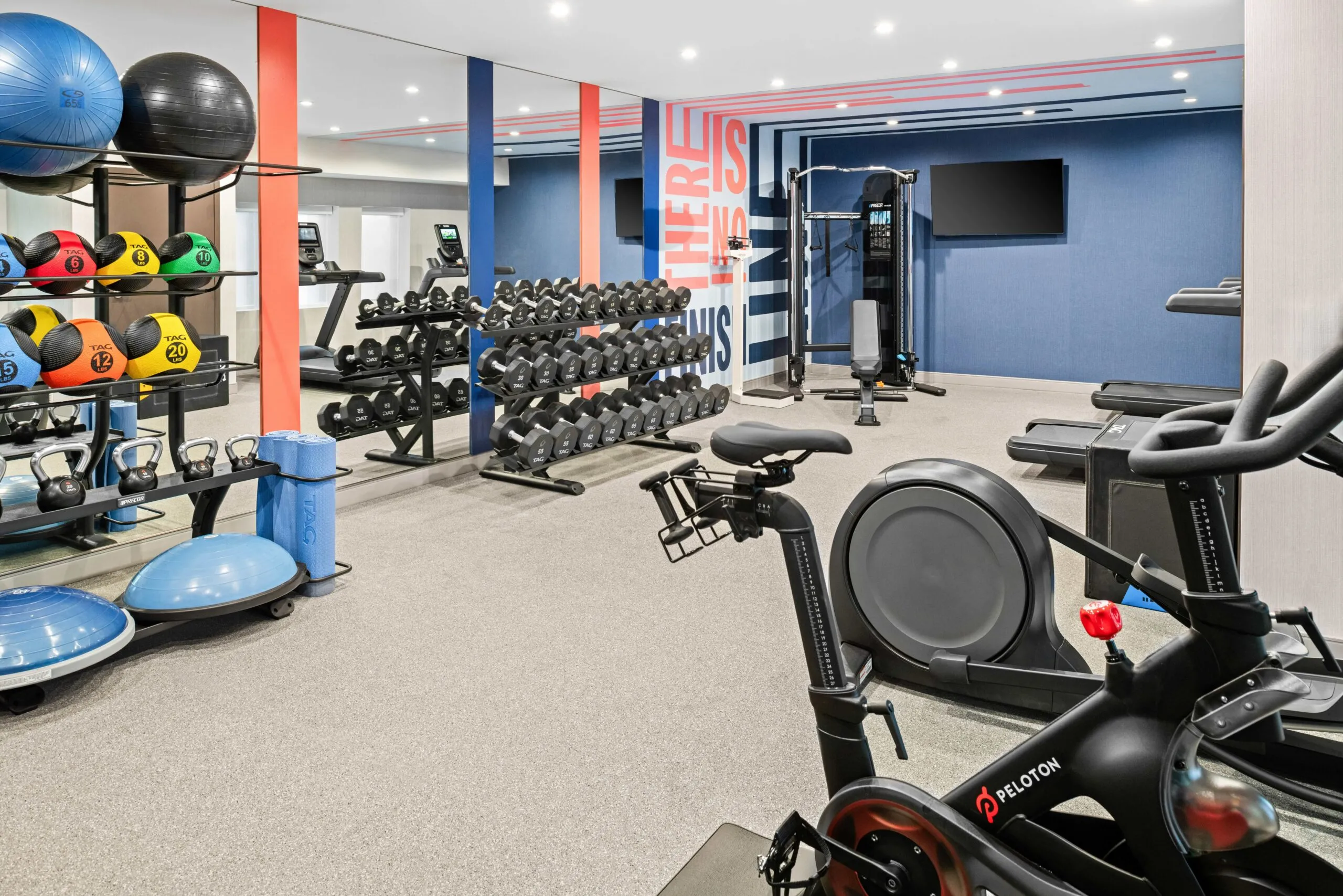 Modern gym at Hampton Inn Costa Mesa Newport Beach with exercise bikes, free weights, dumbbells, kettlebells, medicine balls, mats, BOSU balls, resistance bands, and cardio machines. Blue, gray, and red accents with bright lighting and a wall-mounted TV.