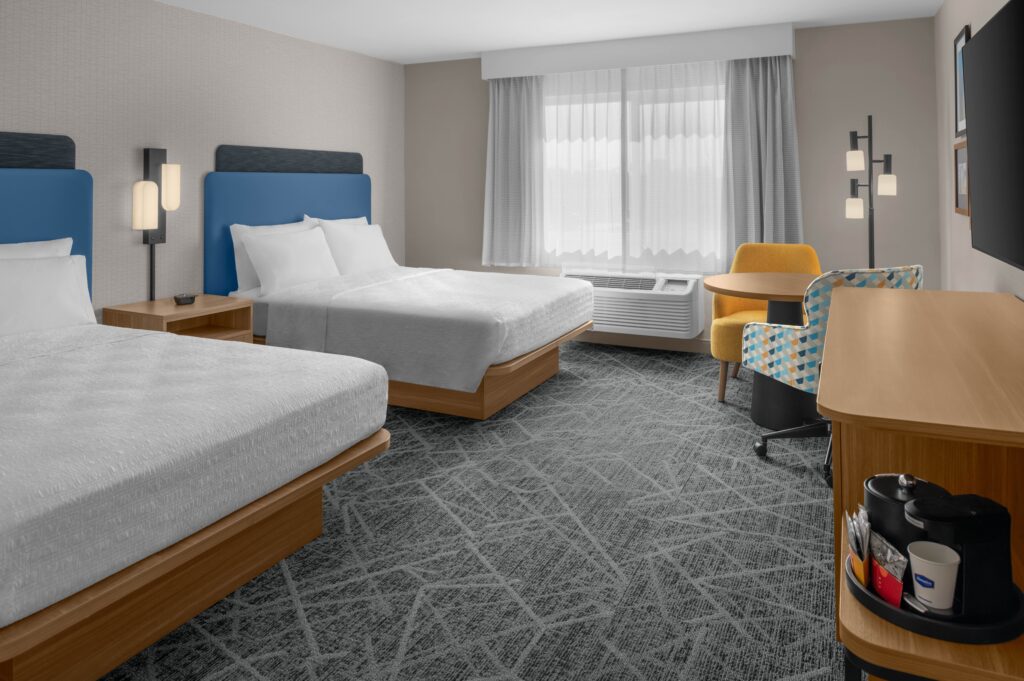 Modern Hampton Inn Costa Mesa Newport Beach hotel room with two double beds, blue headboards, a window with sheer curtains, a yellow chair at a desk, floor lamp, and coffee station on a wooden cabinet. Decor is neutral and contemporary.