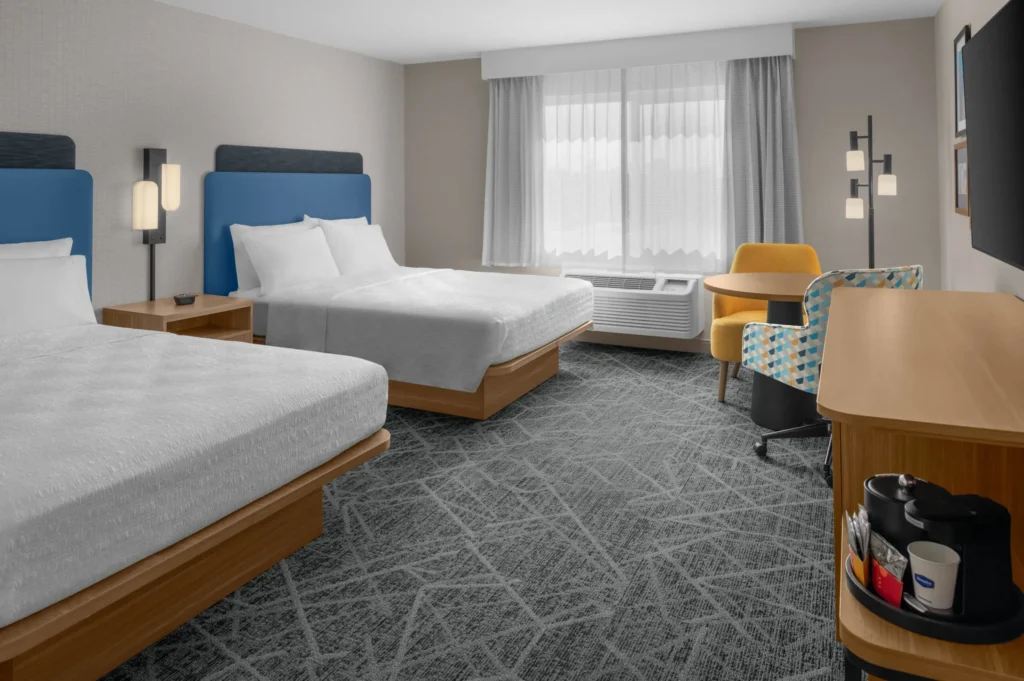 Modern Hampton Inn Costa Mesa Newport Beach hotel room with two double beds, blue headboards, a window with sheer curtains, a yellow chair at a desk, floor lamp, and coffee station on a wooden cabinet. Decor is neutral and contemporary.