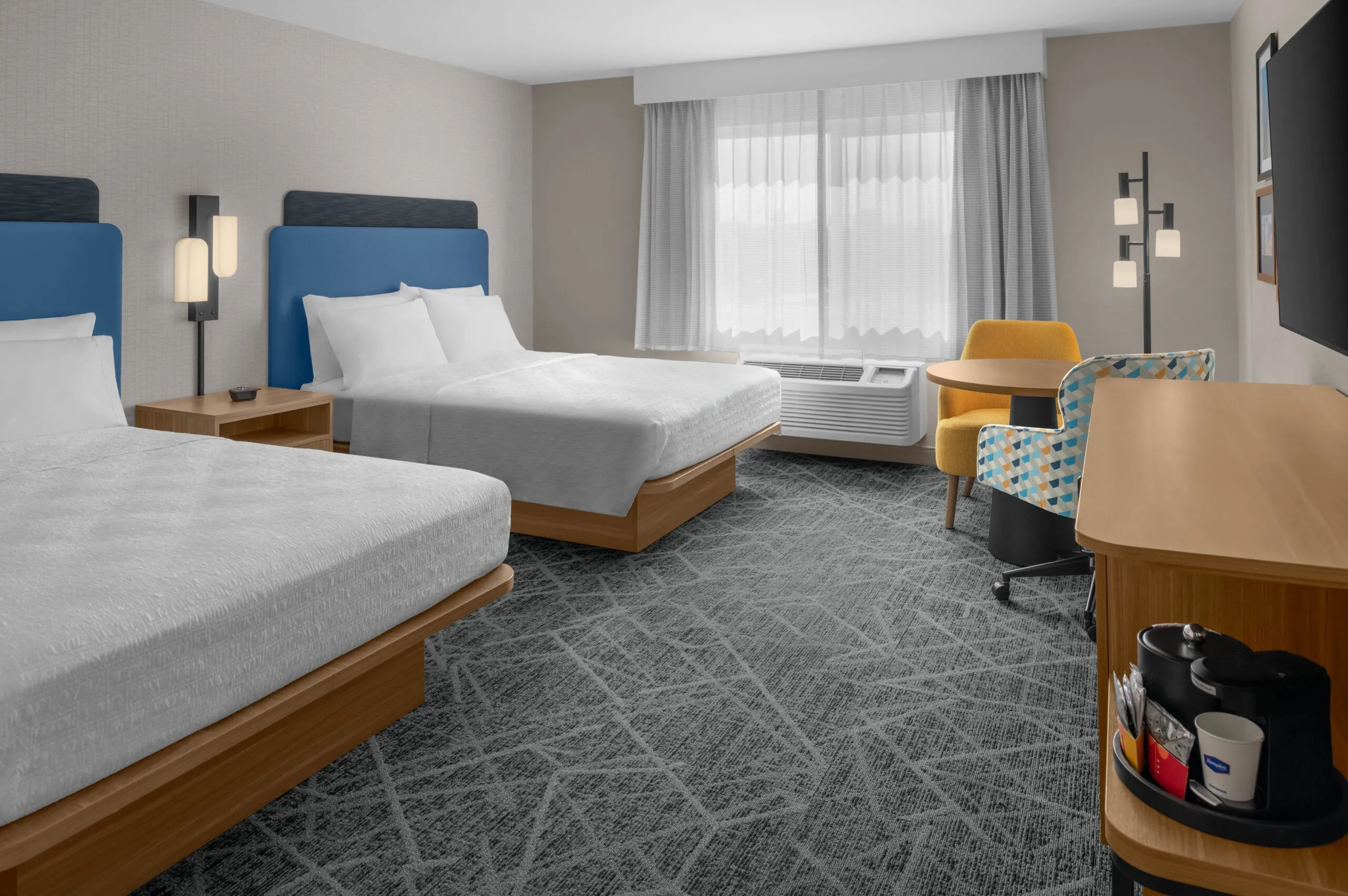 Modern Hampton Inn Costa Mesa Newport Beach hotel room with two double beds, blue headboards, a window with sheer curtains, a yellow chair at a desk, floor lamp, and coffee station on a wooden cabinet. Decor is neutral and contemporary.