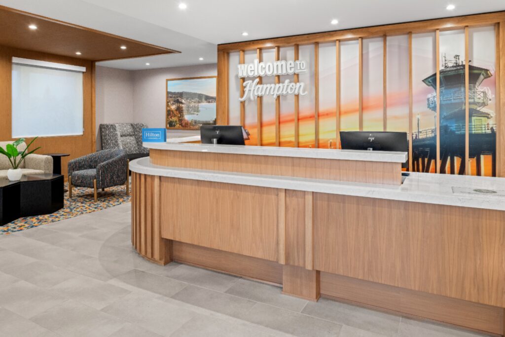 Hotel lobby at Hampton Inn Costa Mesa Newport Beach with a large wooden front desk, modern chairs, two black monitors, a small table with a plant, and a wall mural that says 