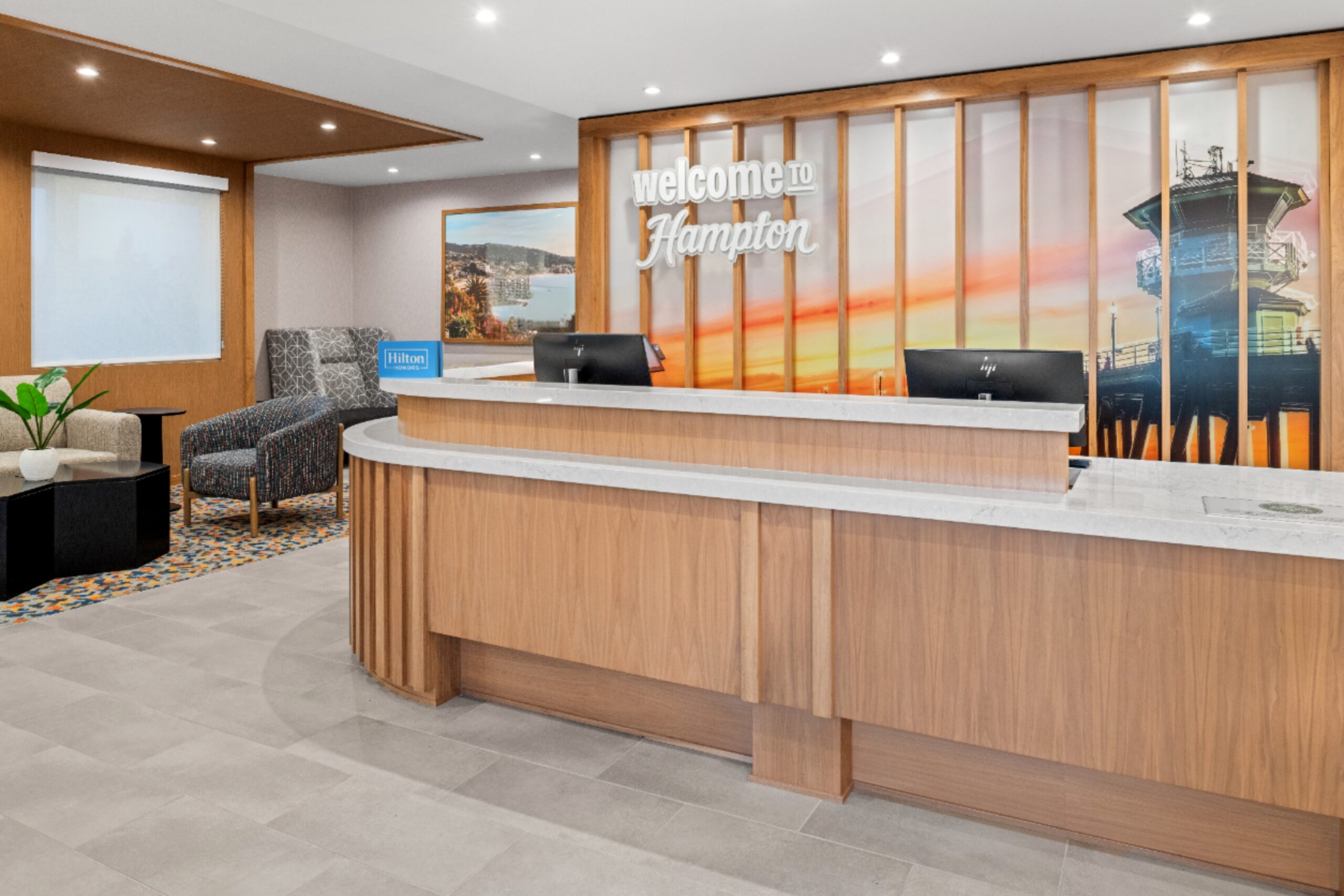 Hotel lobby at Hampton Inn Costa Mesa Newport Beach with a large wooden front desk, modern chairs, two black monitors, a small table with a plant, and a wall mural that says 