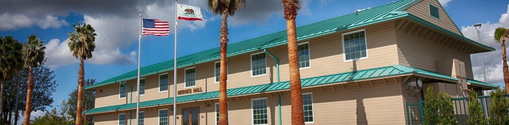 A beige two-story building with a green roof, palm trees in front, and two flags—U.S. and California—flying under a partly cloudy sky. The building sign reads TOWN HALL, reminiscent of the civic style seen at Heroes Hall Museum in Orange County.