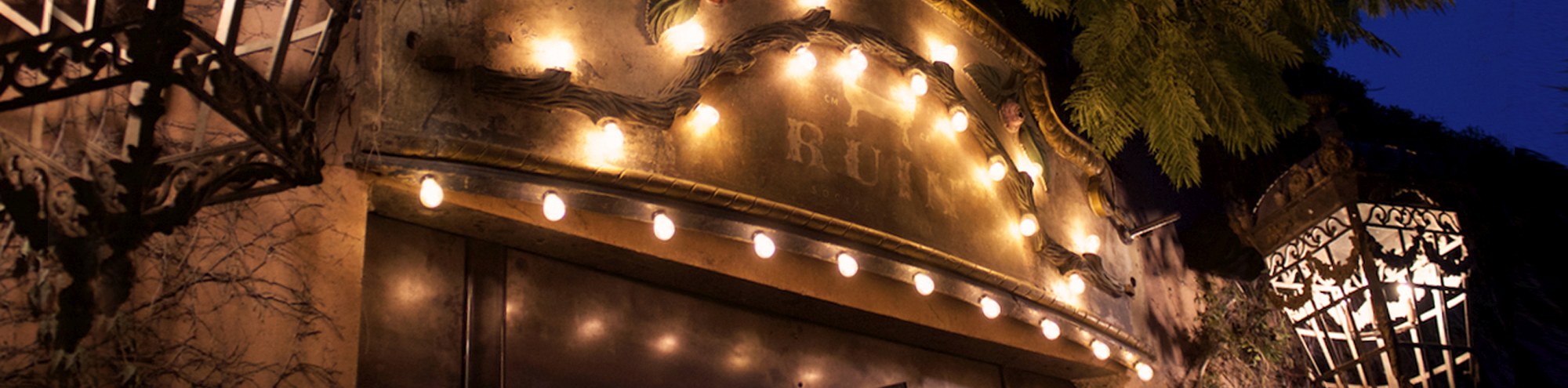 A warmly lit entrance with a rustic sign and string lights, flanked by two ornate lanterns, set against a dark evening sky and leafy greenery.