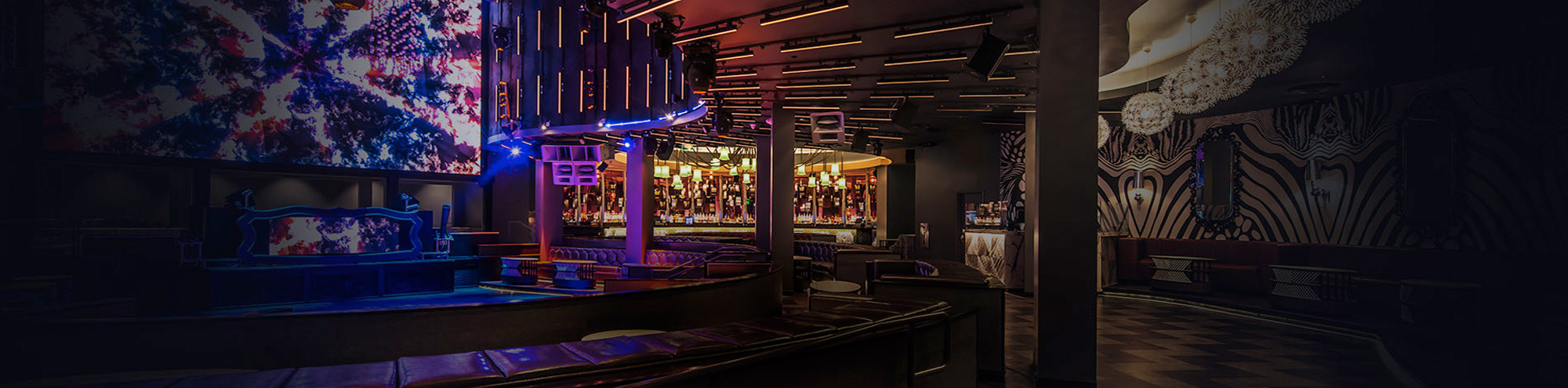 A modern nightclub interior with colorful neon lighting, abstract wall art, a large digital display, cozy leather seating, and a well-lit bar area in the background. The atmosphere is stylish and vibrant.