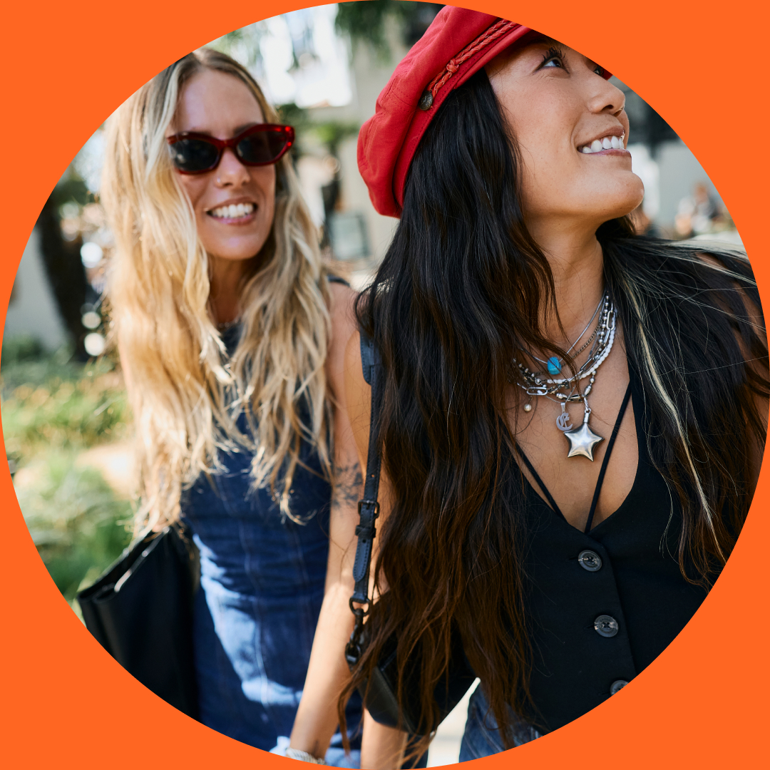 Two women outdoors in Costa Mesa, smiling and walking. One has long blonde hair, sunglasses, and a blue dress. The other has long dark hair, a red hat, layered necklaces, and a black top. The image is framed in an orange circle.