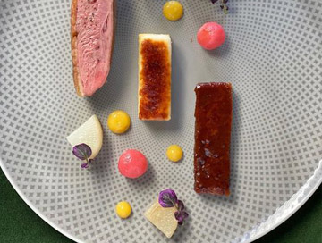 A plated gourmet dish, inspired by Costa Mesa City of the Arts, features slices of seared meat, two rectangular pieces with caramelized tops, colorful sauce dots, and small garnishes on a patterned gray plate.