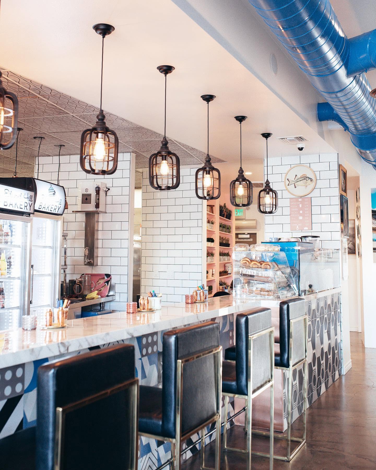 A modern café interior at Tres Muchachos Costa Mesa with a marble countertop, black leather barstools, geometric patterned tiles, hanging cage lights, white subway tile walls, and a blue exposed air duct on the ceiling.