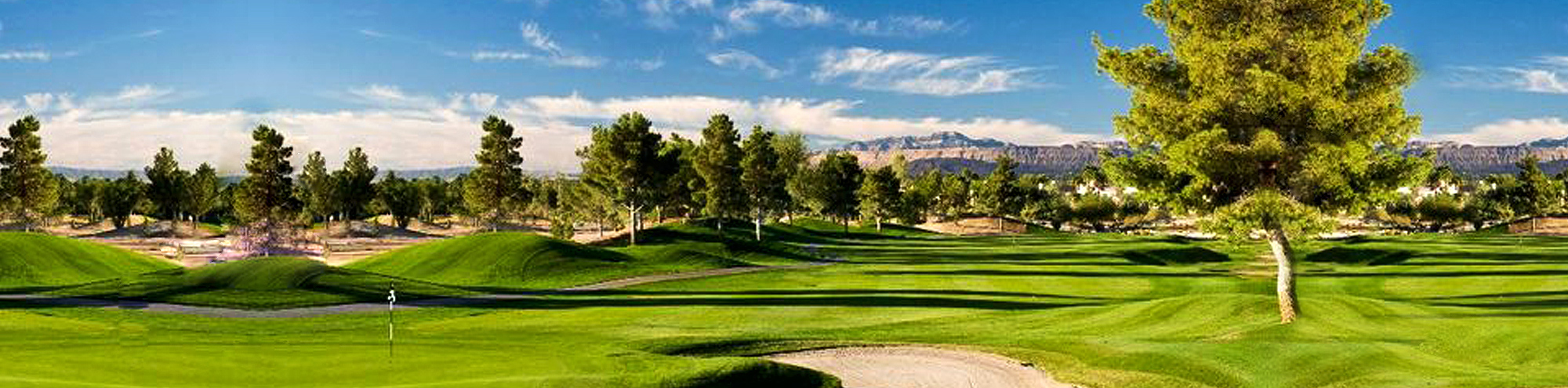 A scenic golf course with lush green fairways, sand bunkers, and tall trees under a bright blue sky, with mountains visible in the background.