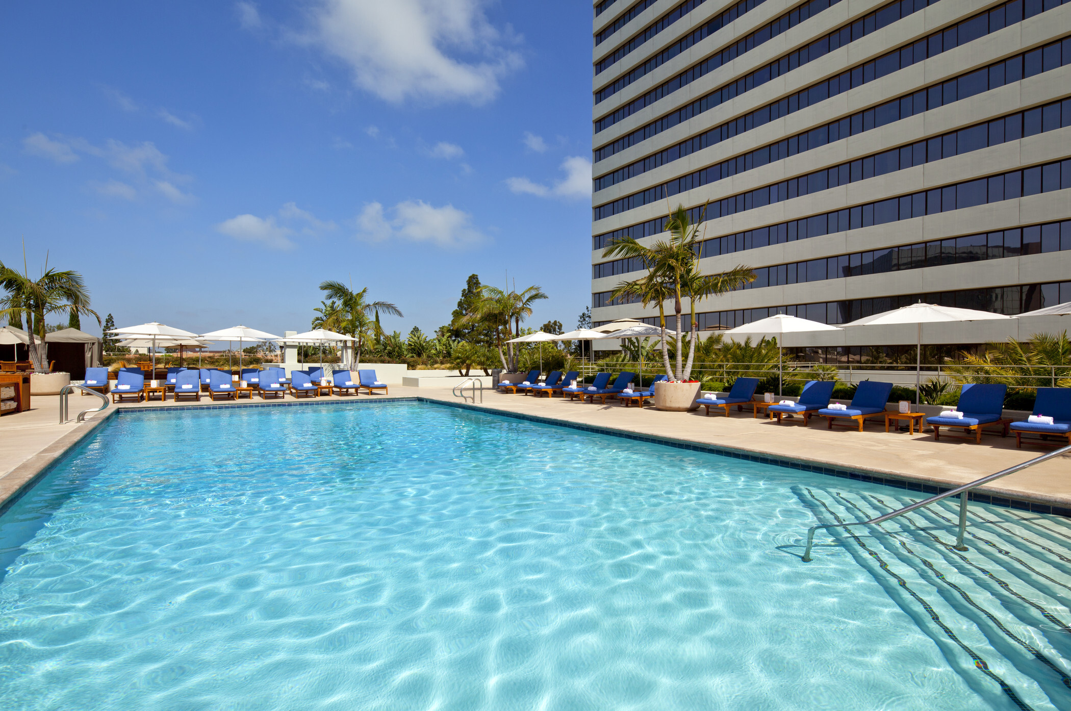 A large outdoor swimming pool surrounded by blue lounge chairs, white umbrellas, palm trees, and a tall modern building under a bright blue sky with scattered clouds—an inviting escape at hotels Costa Mesa.