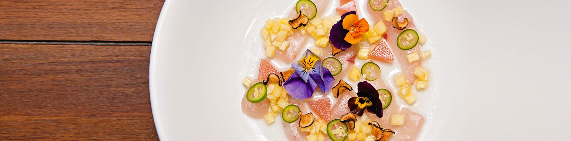 A white plate with artfully arranged pieces of raw fish, thinly sliced green chilies, diced yellow fruit, toasted seeds, and colorful edible flowers on a wooden table.
