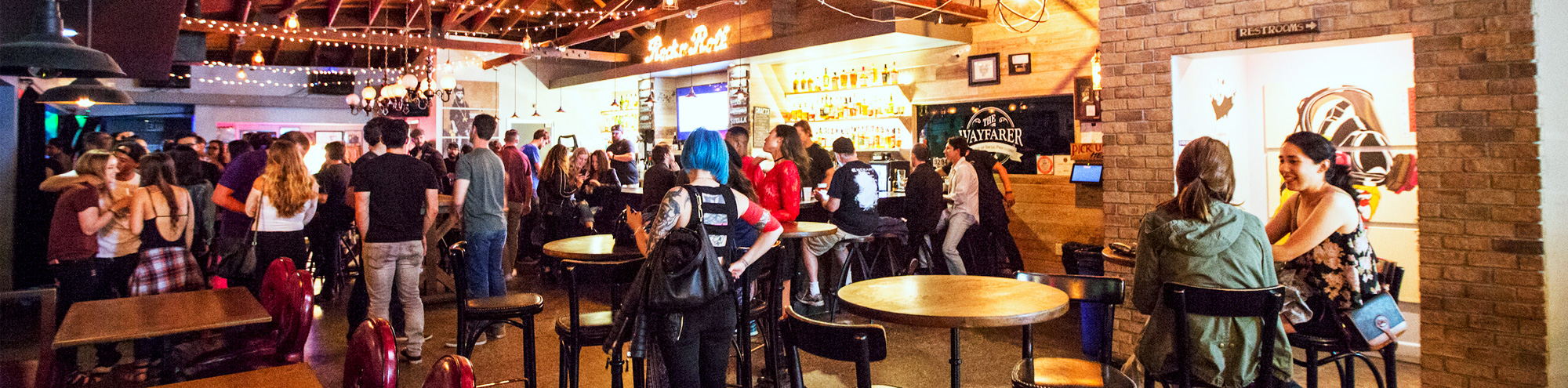 A lively bar scene with groups of people standing and sitting at tables, colorful lights strung across the ceiling, a wooden bar area, and a bright, welcoming atmosphere.