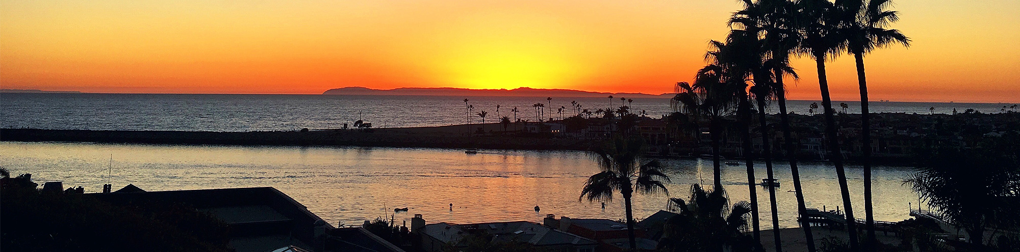 A vibrant orange sunset over the ocean at Corona del Mar Beach, Orange County, with an island in the distance, silhouetted palm trees, and a calm body of water reflecting the sky. Houses and buildings line the shore.