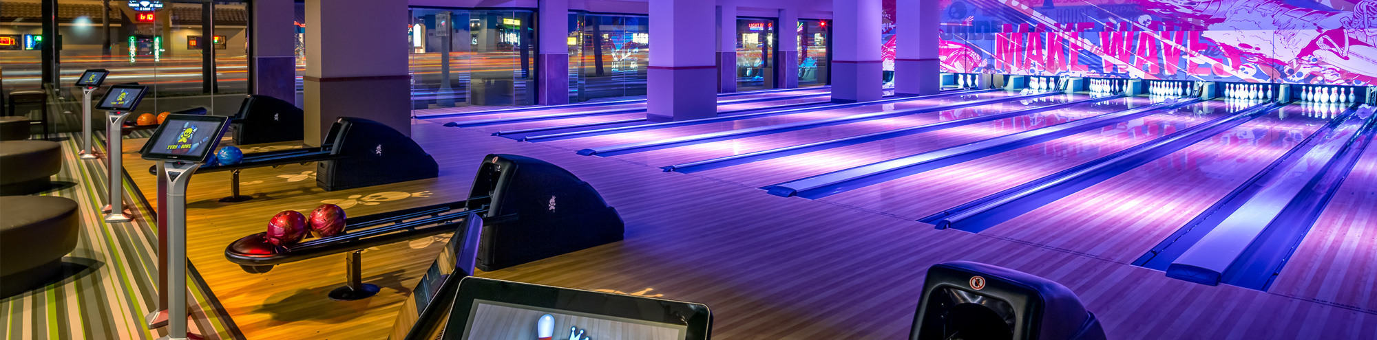 A modern, brightly lit bowling alley with multiple empty lanes, colorful bowling balls, and illuminated lane markings. Large windows reveal blurred city lights outside.