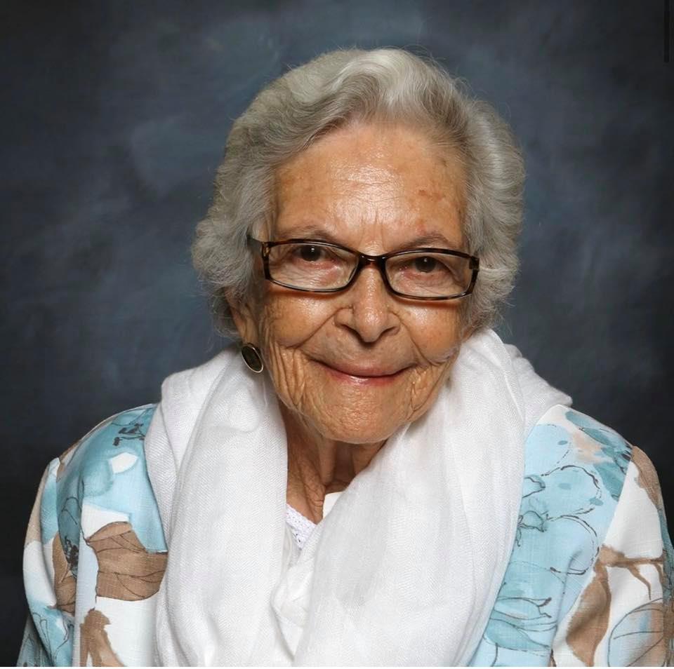 An elderly woman with white hair, glasses, and a gentle smile wears a white scarf and a light blue floral top, posed against a dark, neutral background.