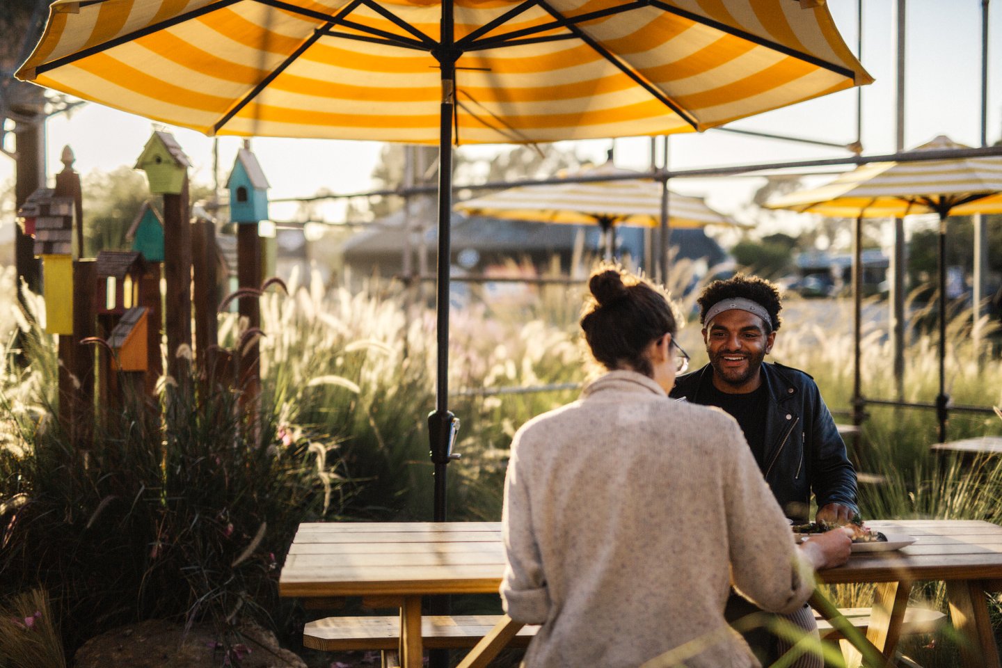 Two people sit across from each other at a wooden picnic table outdoors, under a large yellow-striped umbrella, with plants, decorative birdhouses, and sunlight in the background.