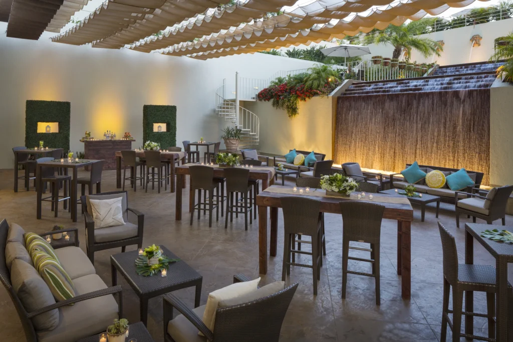 A stylish outdoor patio with high tables and chairs, cushioned sofas, decorative plants, and a large water feature on one wall. Ideal for gatherings or as one of the most inviting wedding venues Costa Mesa offers, with a shaded canopy creating a cozy atmosphere.