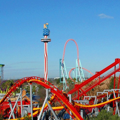A theme park with colorful roller coasters, including a red and yellow track in the foreground and a tall tower decorated with stars and stripes under a clear blue sky.
