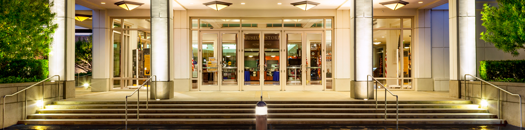 Well-lit building entrance at night, featuring wide glass doors, illuminated columns, stairs with metal railings, and greenery on both sides. The interior appears to be a gift shop or store.