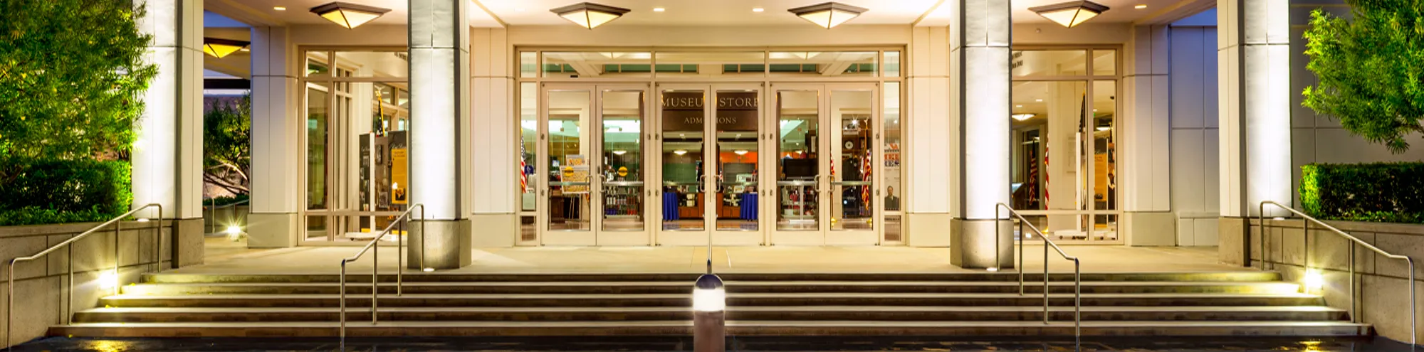 Well-lit building entrance at night, featuring wide glass doors, illuminated columns, stairs with metal railings, and greenery on both sides. The interior appears to be a gift shop or store.